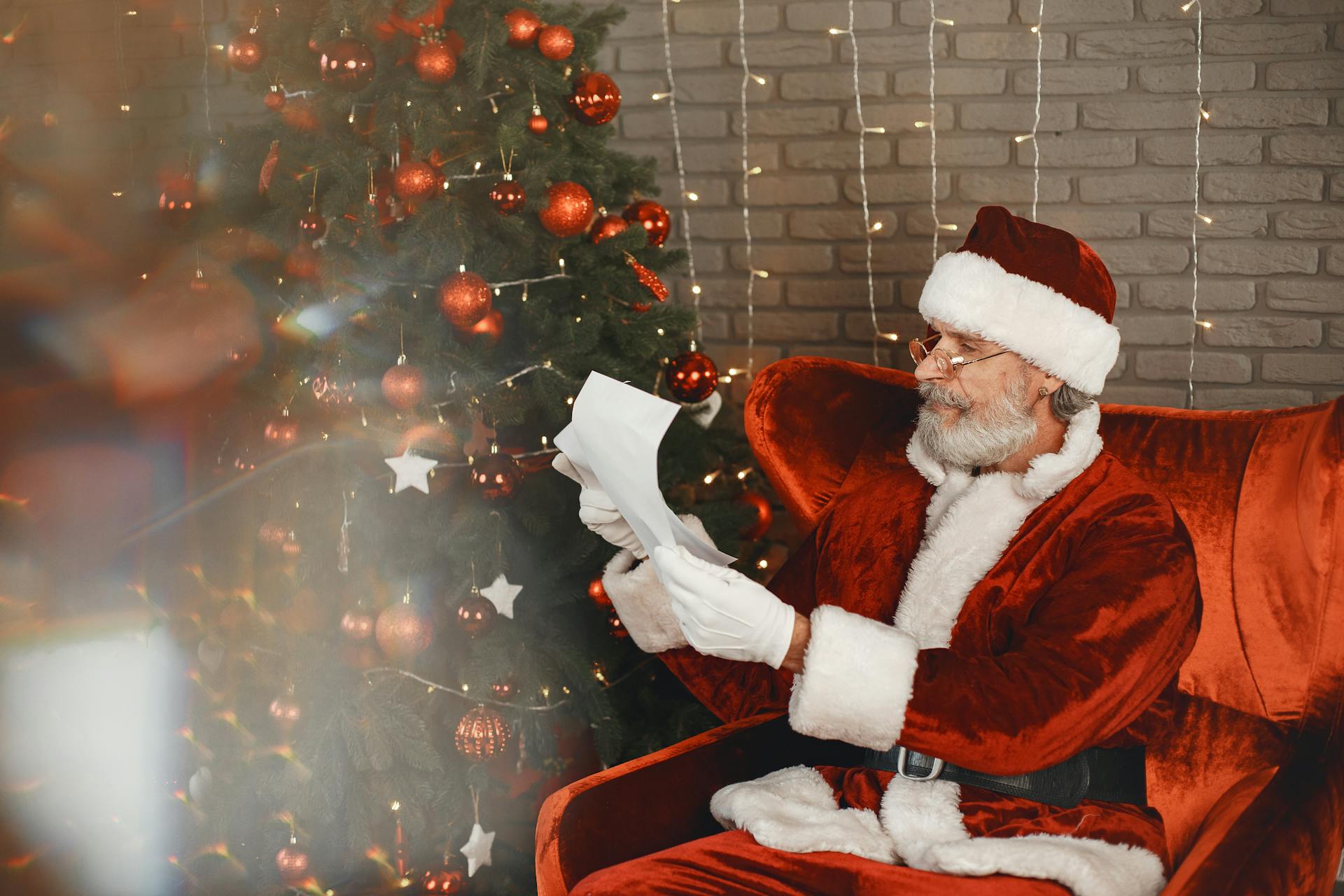 Santa Claus reading a letter in his red costume next to a decorated Christmas tree.