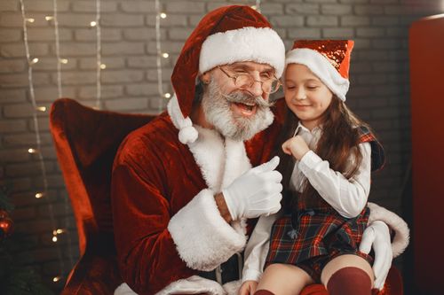 Free Santa Claus shares a joyful moment with a child wearing a red hat and plaid dress. Stock Photo