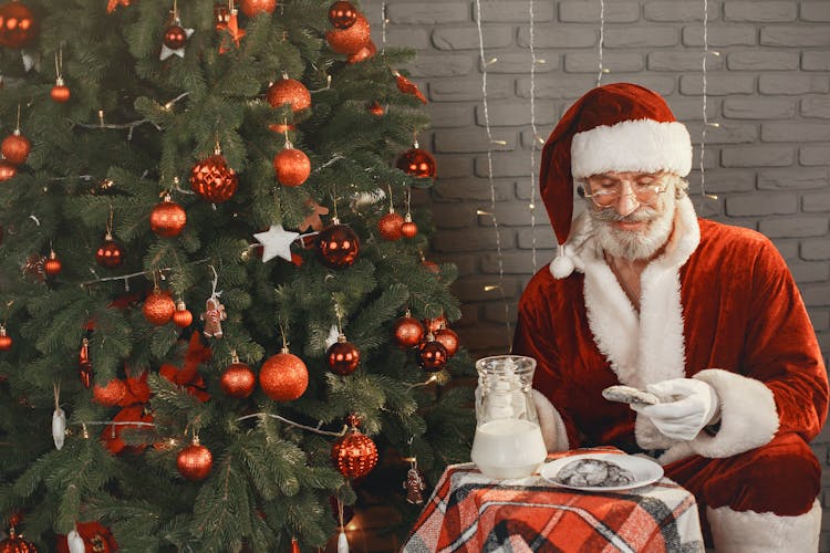 Man Dressed As Santa Clause Sitting Next To A Christmas Tree And Eating Cookies 