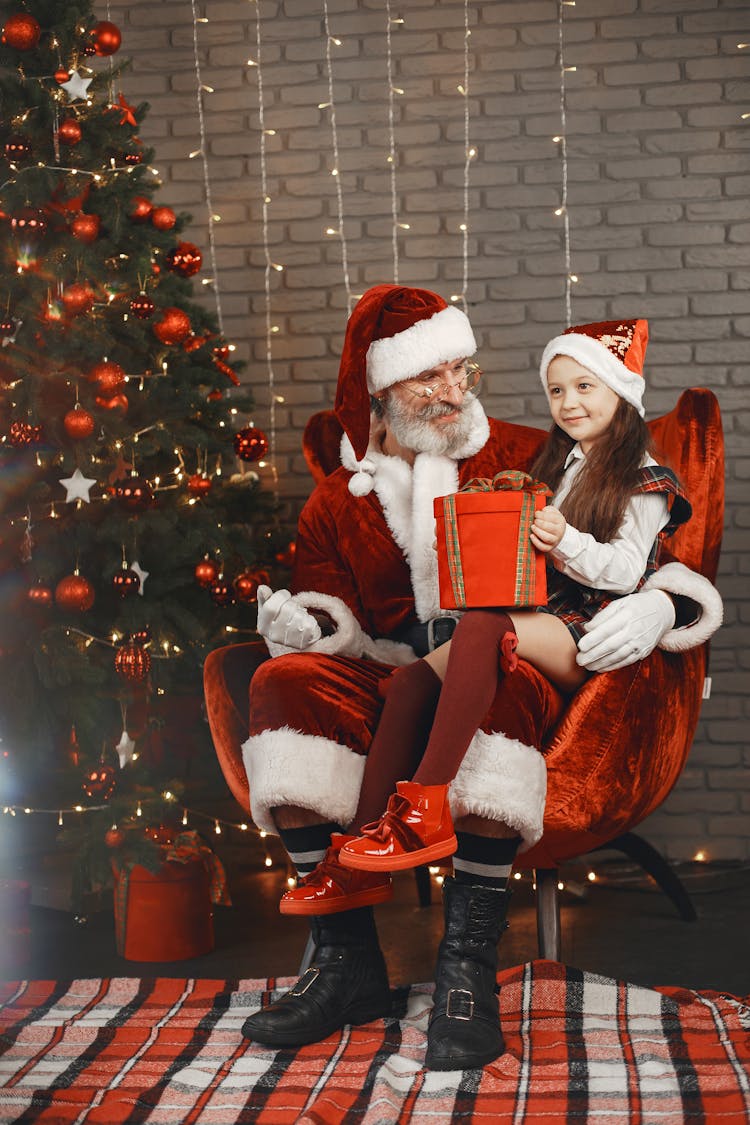 Girl With A Christmas Gift Sitting On The Lap Of Santa Claus