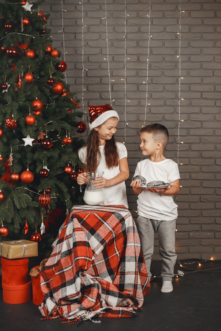 Girl In Santa Hat With A Pitcher Of Milk And Boy Holding A Plate Of Cookies Standing Beside A Christmas Tree