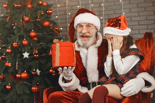 Santa Claus Sitting on Red Sofa Chair Holding a Gift Beside a Girl