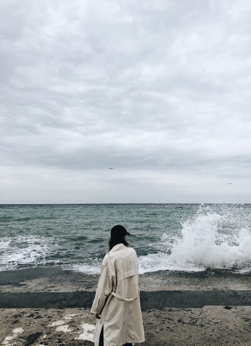 Free Back view of unrecognizable female tourist in stylish coat and cap standing on wet rocky beach and admiring powerful stormy ocean against overcast sky Stock Photo