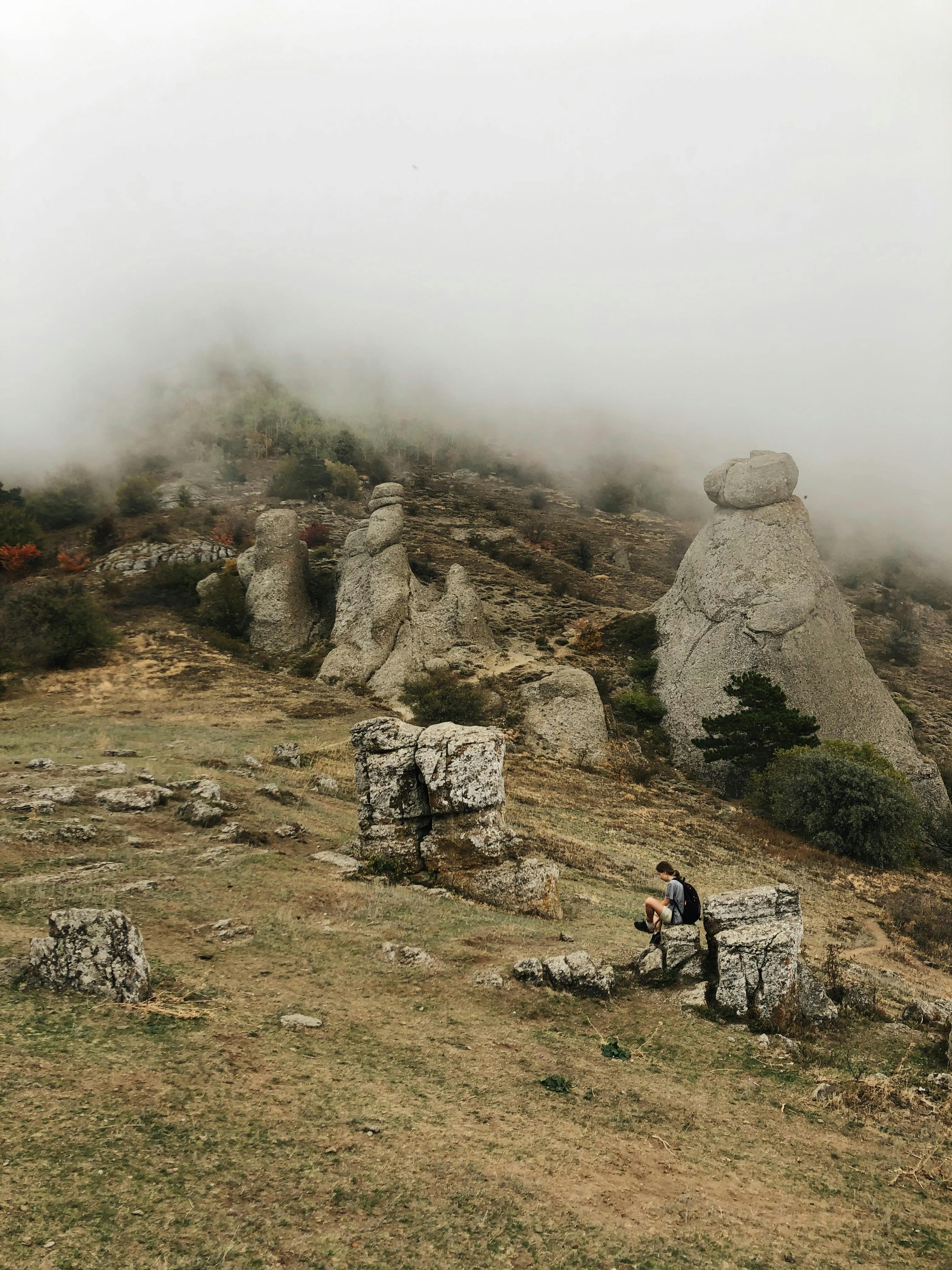anonymous traveler resting on boulder in rocky mountainous terrain against misty sky