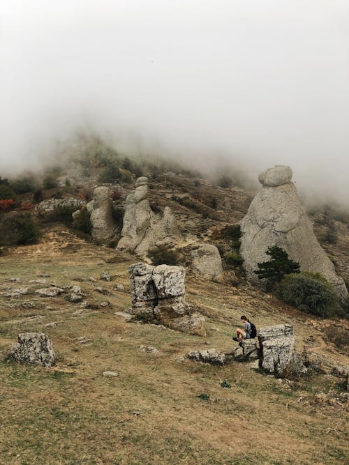Side view of unrecognizable hiker relaxing on rough stone and admiring picturesque view of Valley of Ghosts with various rocky formations against foggy sky in Crimea