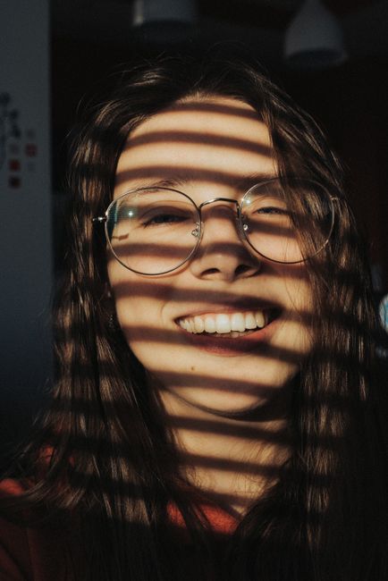 Happy young female in eyeglasses with shadow on face smiling and looking at camera while standing in room with bright sunlight