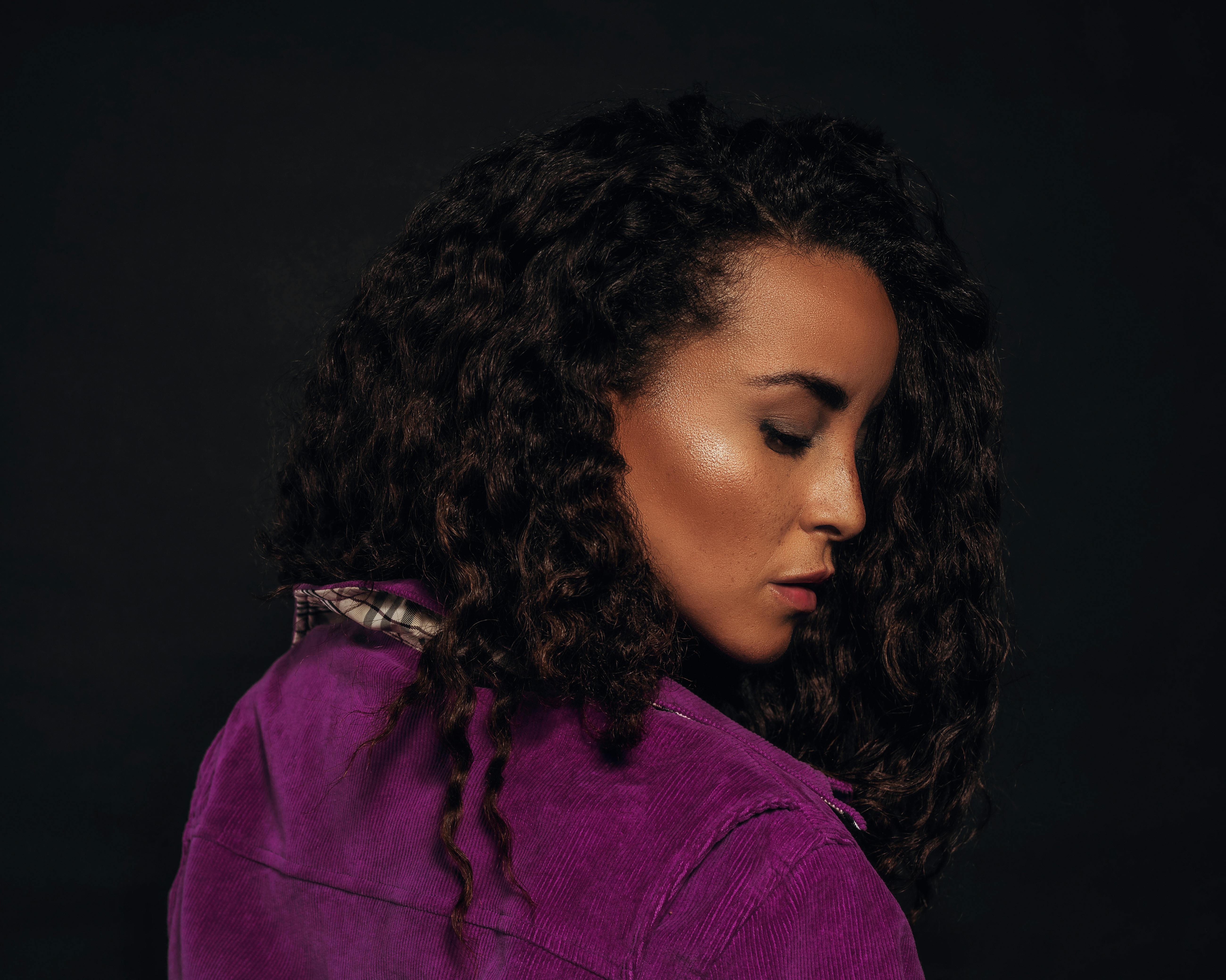 Profile portrait of a woman with curly hair in a purple jacket against a black background.