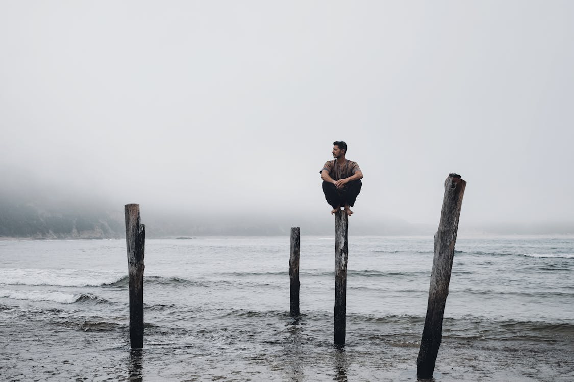 Free Serious male sitting on top of wooden pillar on coast with waving stormy sea against misty sky in overcast weather in nature Stock Photo