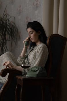 A woman in vintage attire sitting indoors, speaking on a rotary phone.
