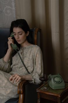Woman seated by a side table with a rotary phone and wine, exuding a nostalgic vintage vibe.