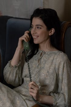 A woman in a vintage dress talks on a retro telephone, smiling indoors.