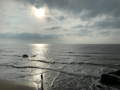 A peaceful beach scene with a lone person standing by the shoreline under a cloudy sky.