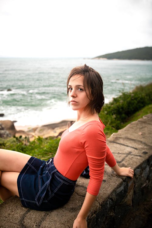 Side view of pensive young lady in casual outfit resting on stone border over rocky beach of wavy ocean and looking at camera during holidays