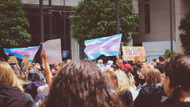 Crowd gathers for a peaceful protest with transgender and Black Lives Matter flags in the street.