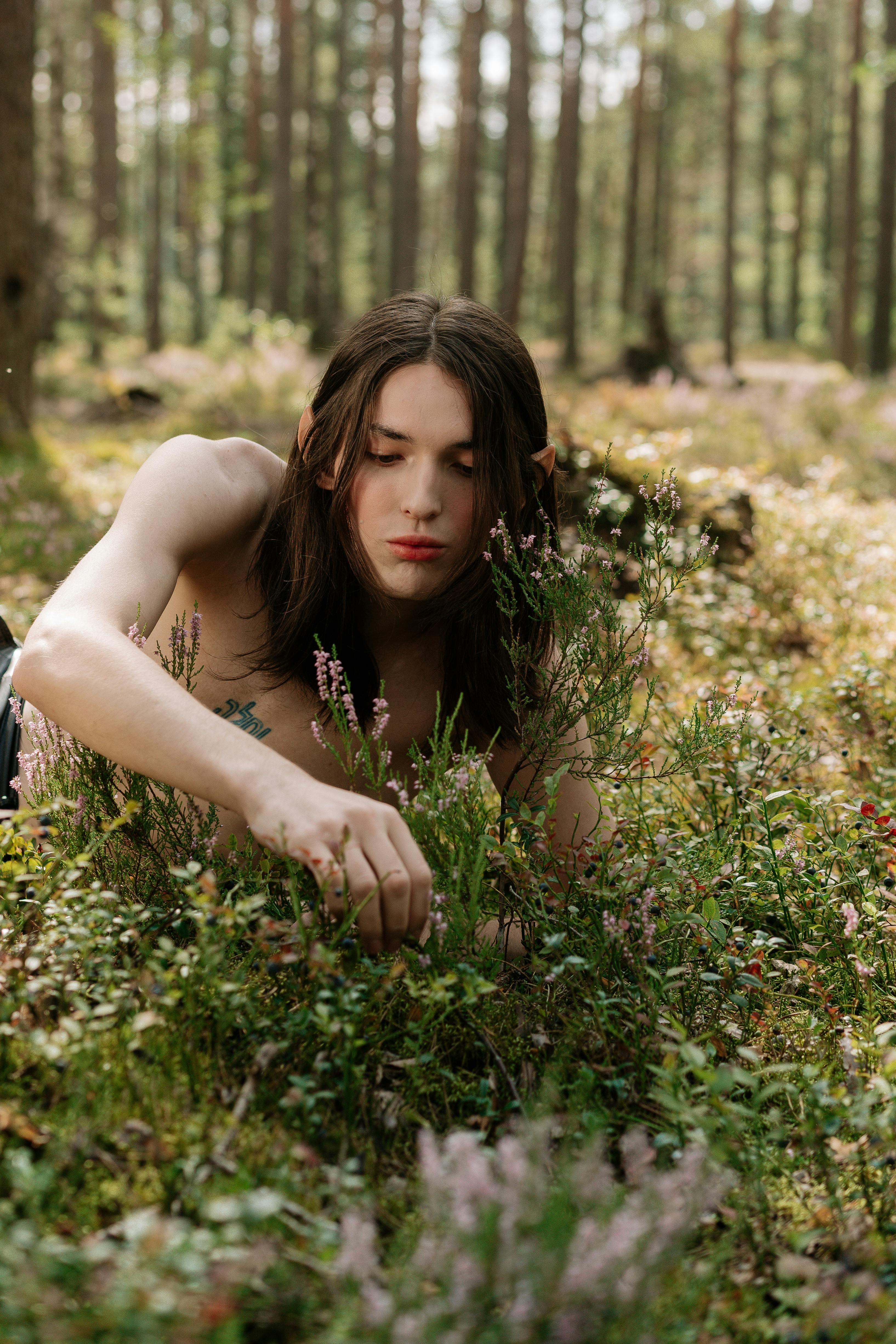 A shirtless man with long hair explores a mystical forest, surrounded by lush greenery and flowers.
