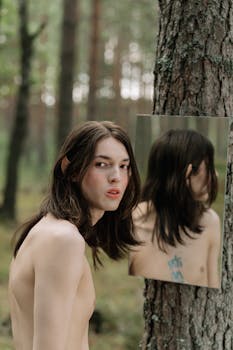 Shirtless person with long hair reflects in a mirror against a forest backdrop.