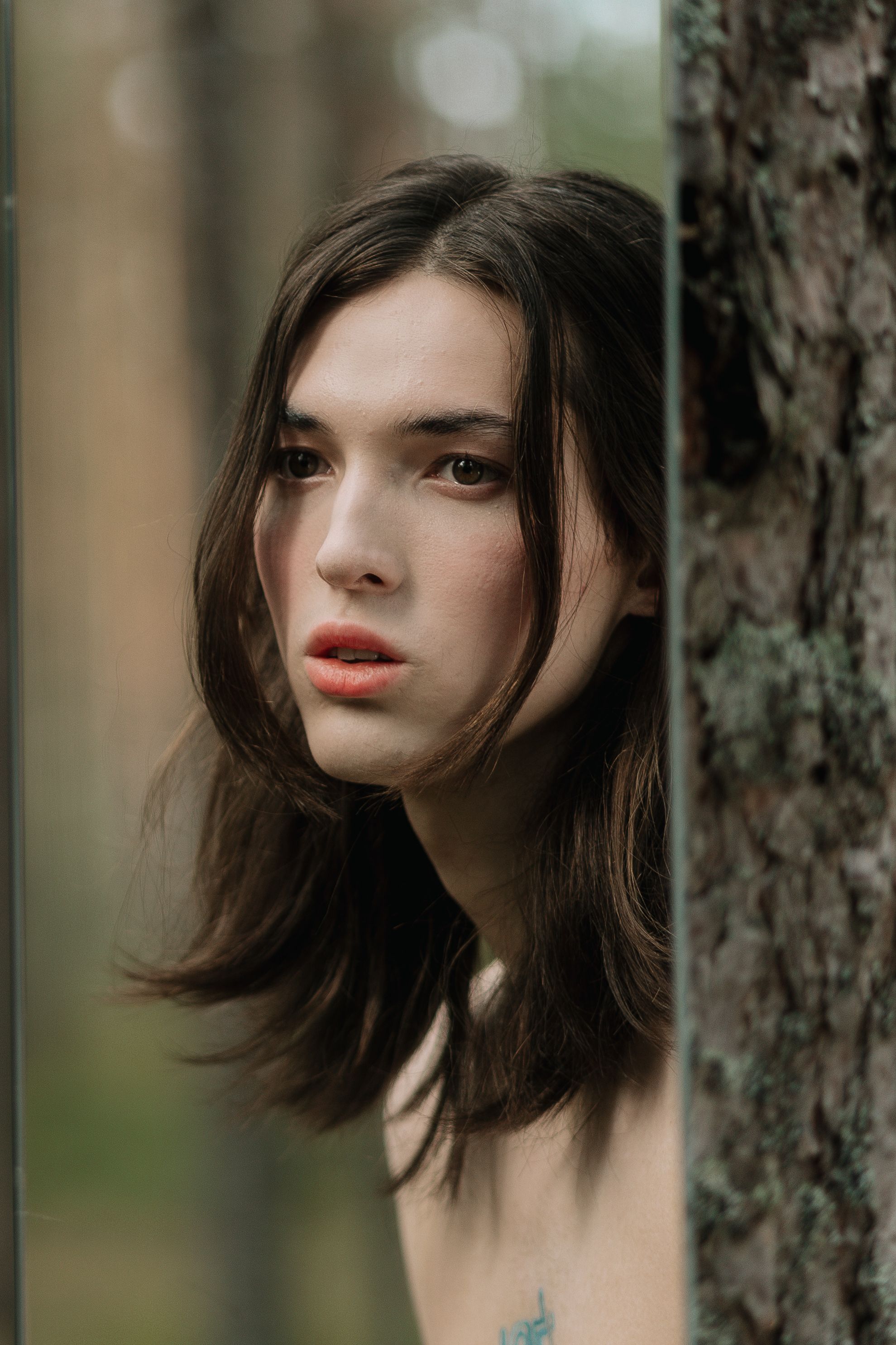 A woman peers around a reflective surface with a mysterious expression in a wooded area.