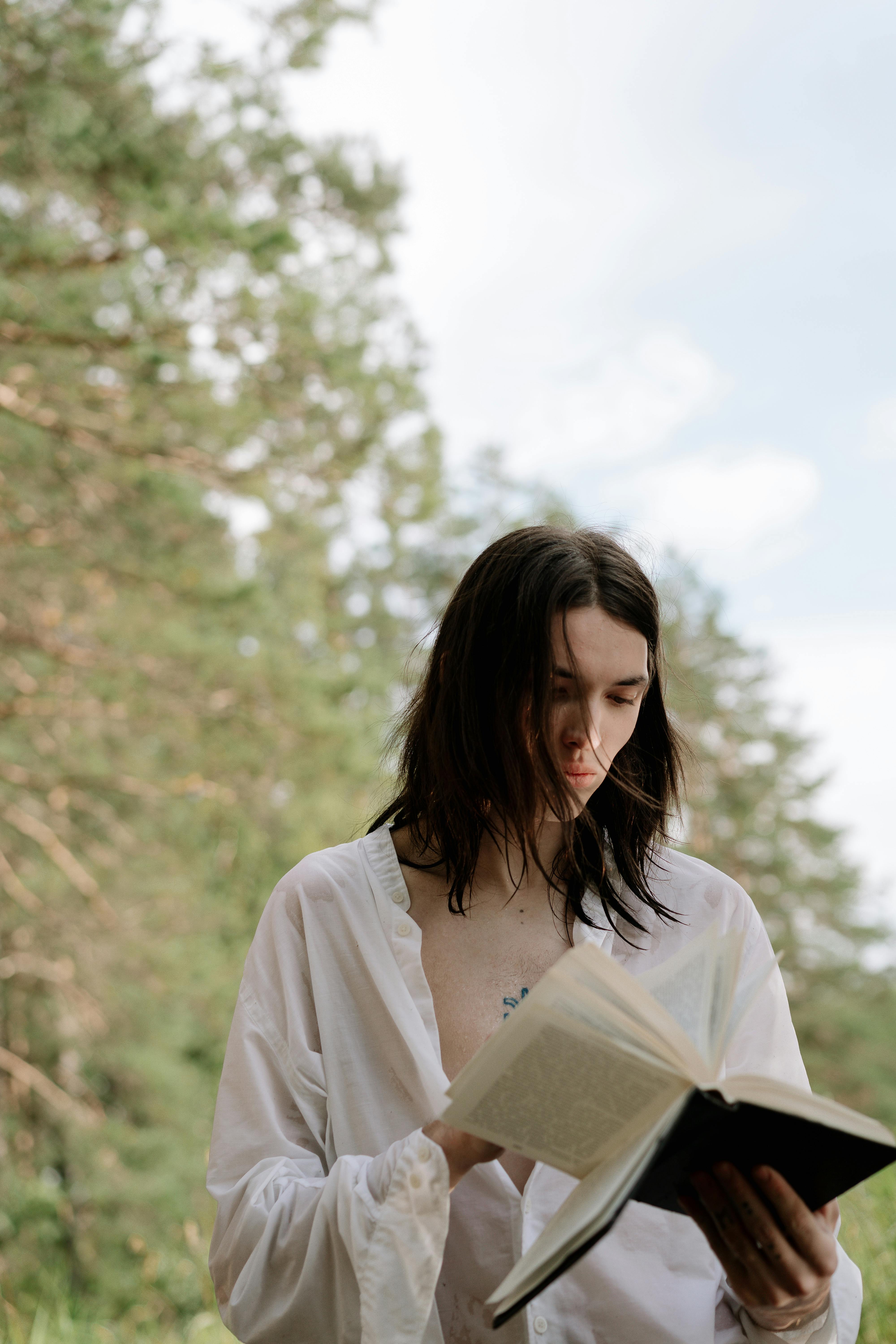 Young adult with long hair reading a book outdoors, surrounded by nature.