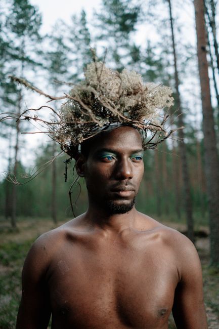 Shirtless man with a unique natural headdress stands in a forest setting, exuding a serious expression.