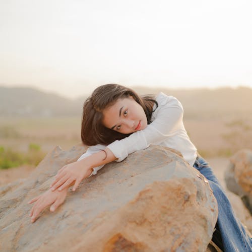 Woman in White Long Sleeves Shirt Lying on Brown Rock