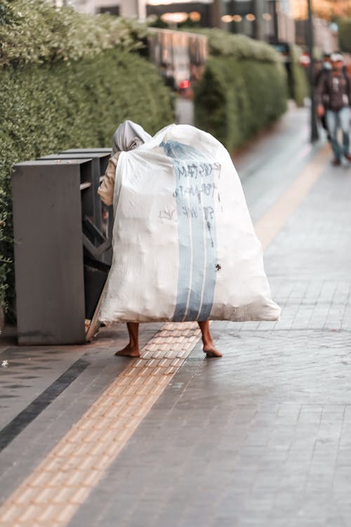 A Barefooted Man Carrying Trash Bag while Walking on the Street