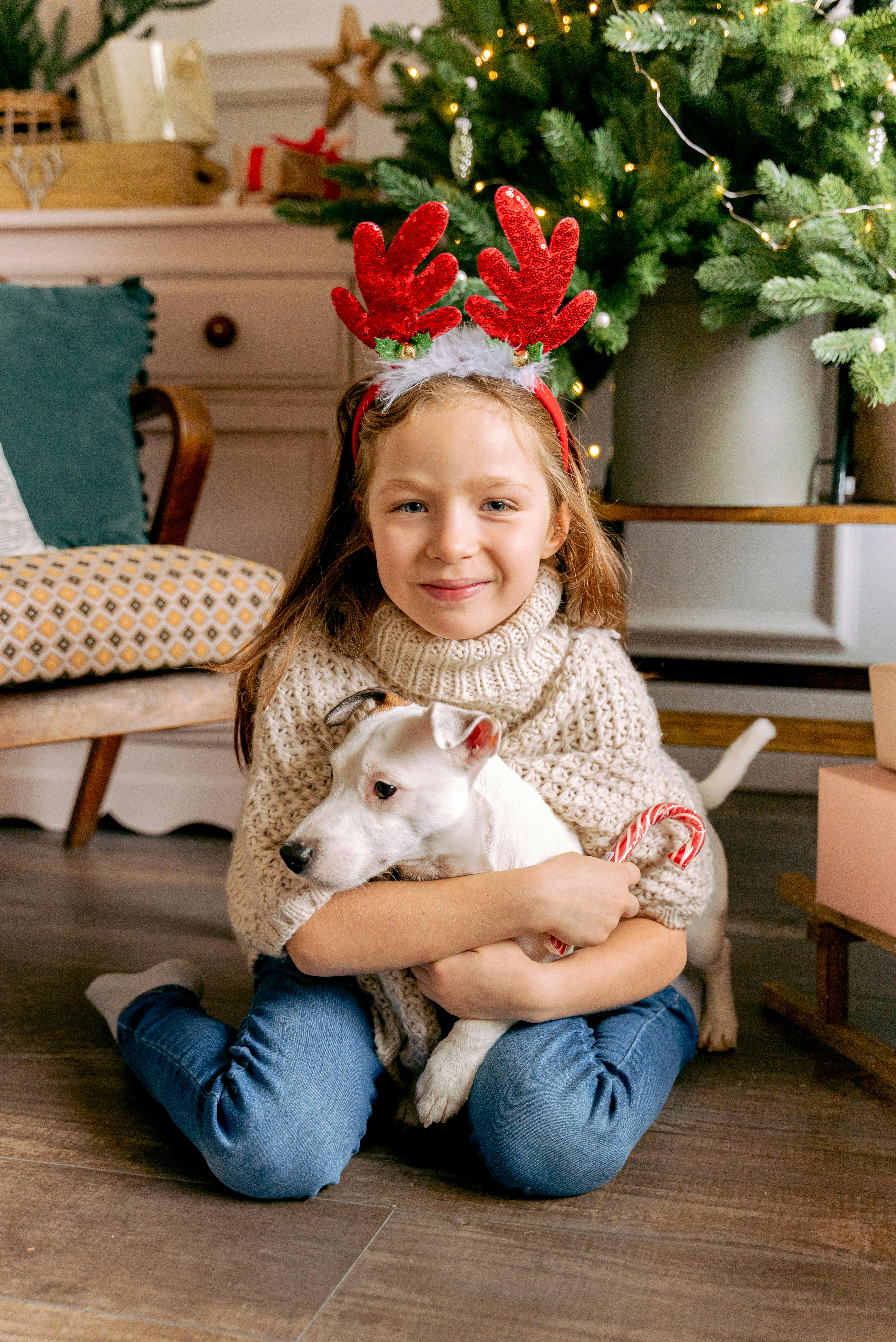 Smiling girl with reindeer headband hugging dog by Christmas tree indoors.