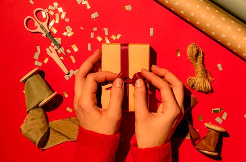 Free Hands wrapping a gift box with ribbon on a vibrant red background, surrounded by festive holiday decorations. Stock Photo