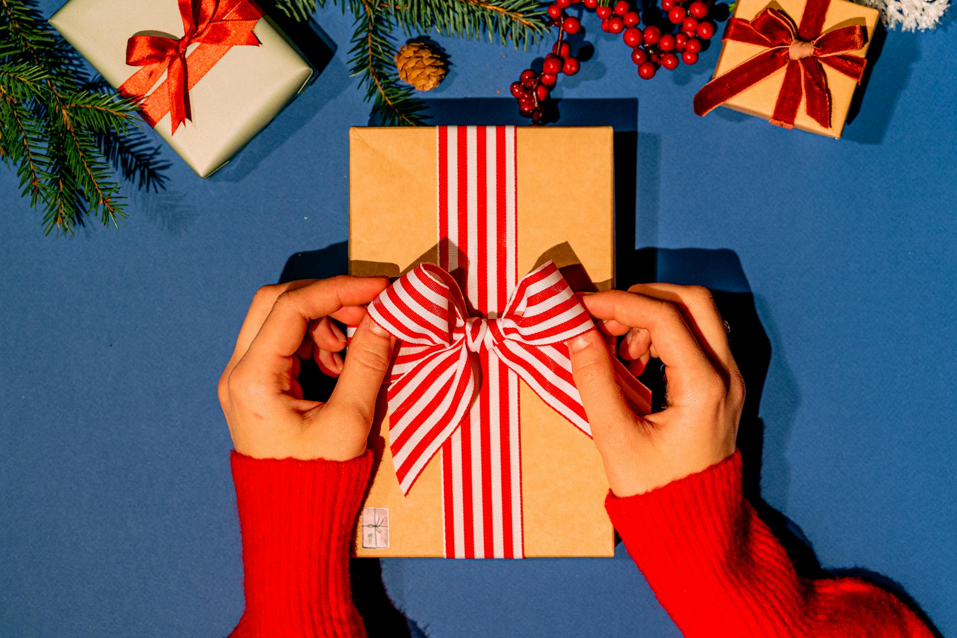 Hands tying a red-striped ribbon on a Christmas gift box on a blue background, surrounded by festive decor.