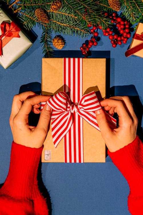 Hands tying a red and white striped ribbon on a gift box surrounded by holiday decorations.