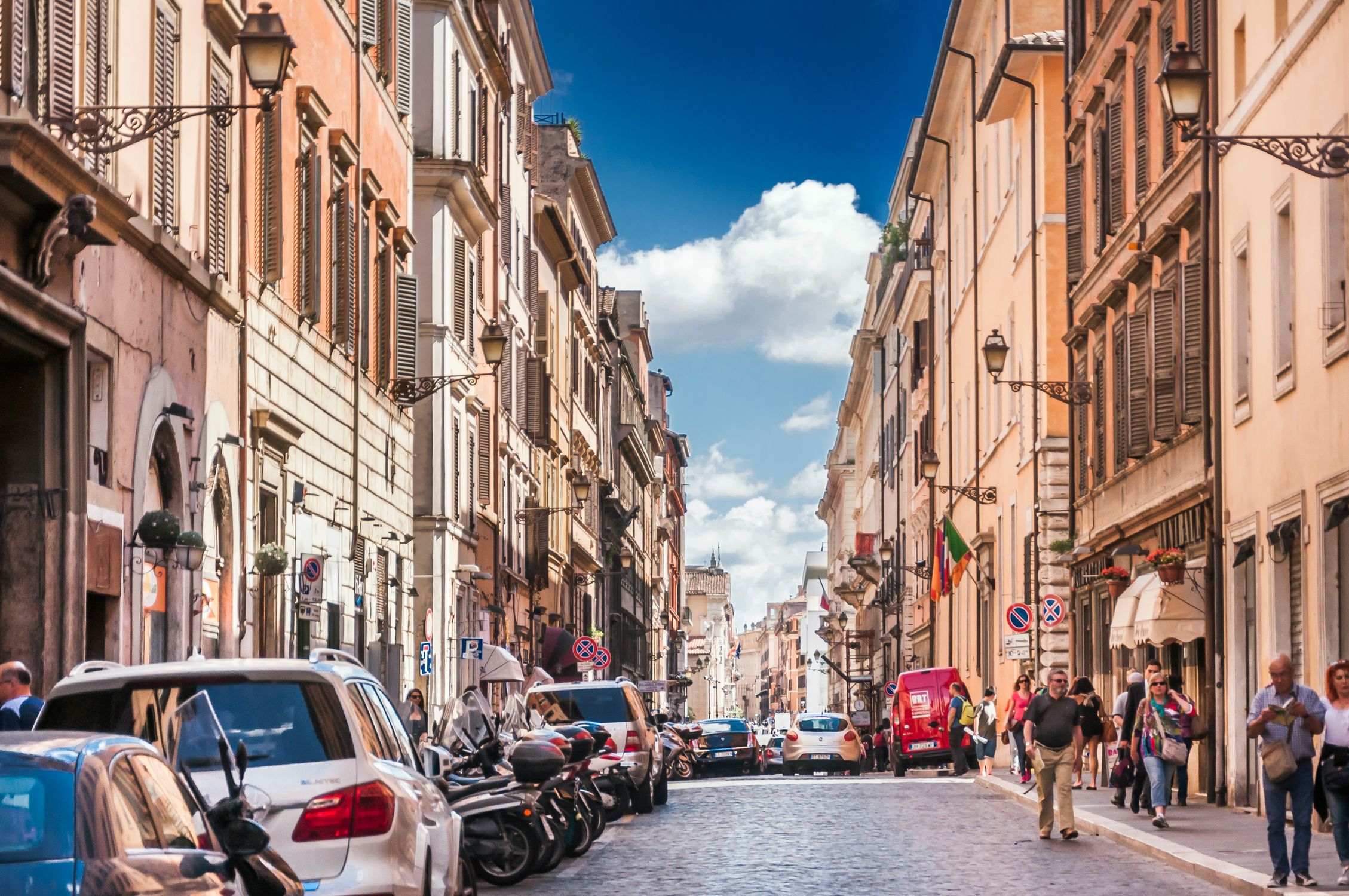 Narrow street with cars and motorcycles parked near historic buildings located in city in Europe and with people walking outside under blue sky in summer sunny day