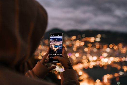 From behind crop person in hoodie taking picture on mobile phone of scenic night city located on hillside and illuminated by bright electric lights