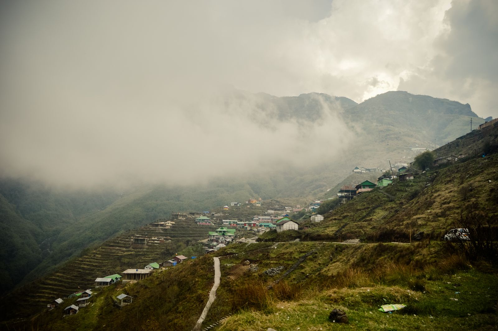 A Sikkim hill village on terraced slopes with cloud pouring over the ridge above it and a track winding through