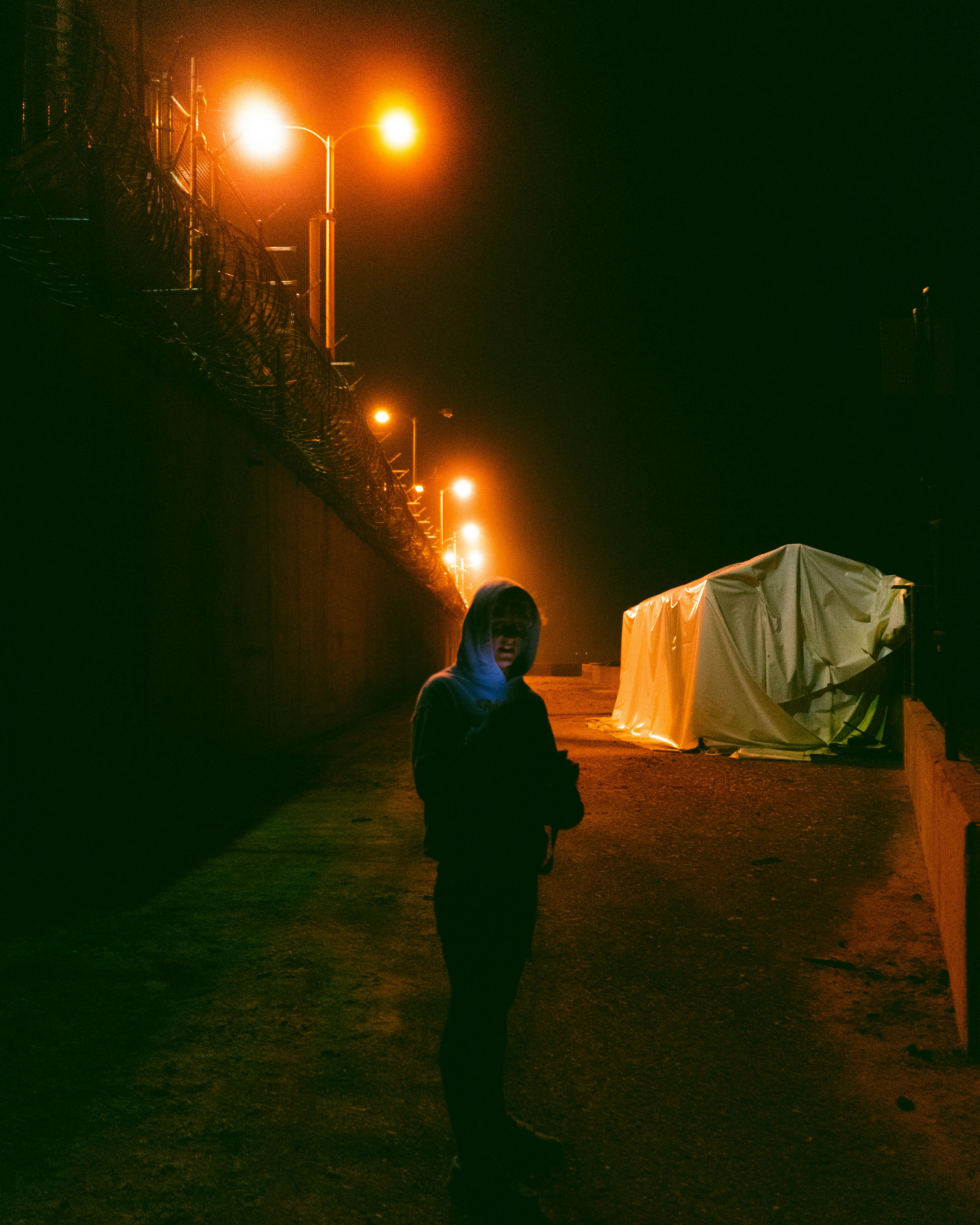 Full body of unrecognizable person standing on walkway near concrete wall on street with glowing streetlight at night in city