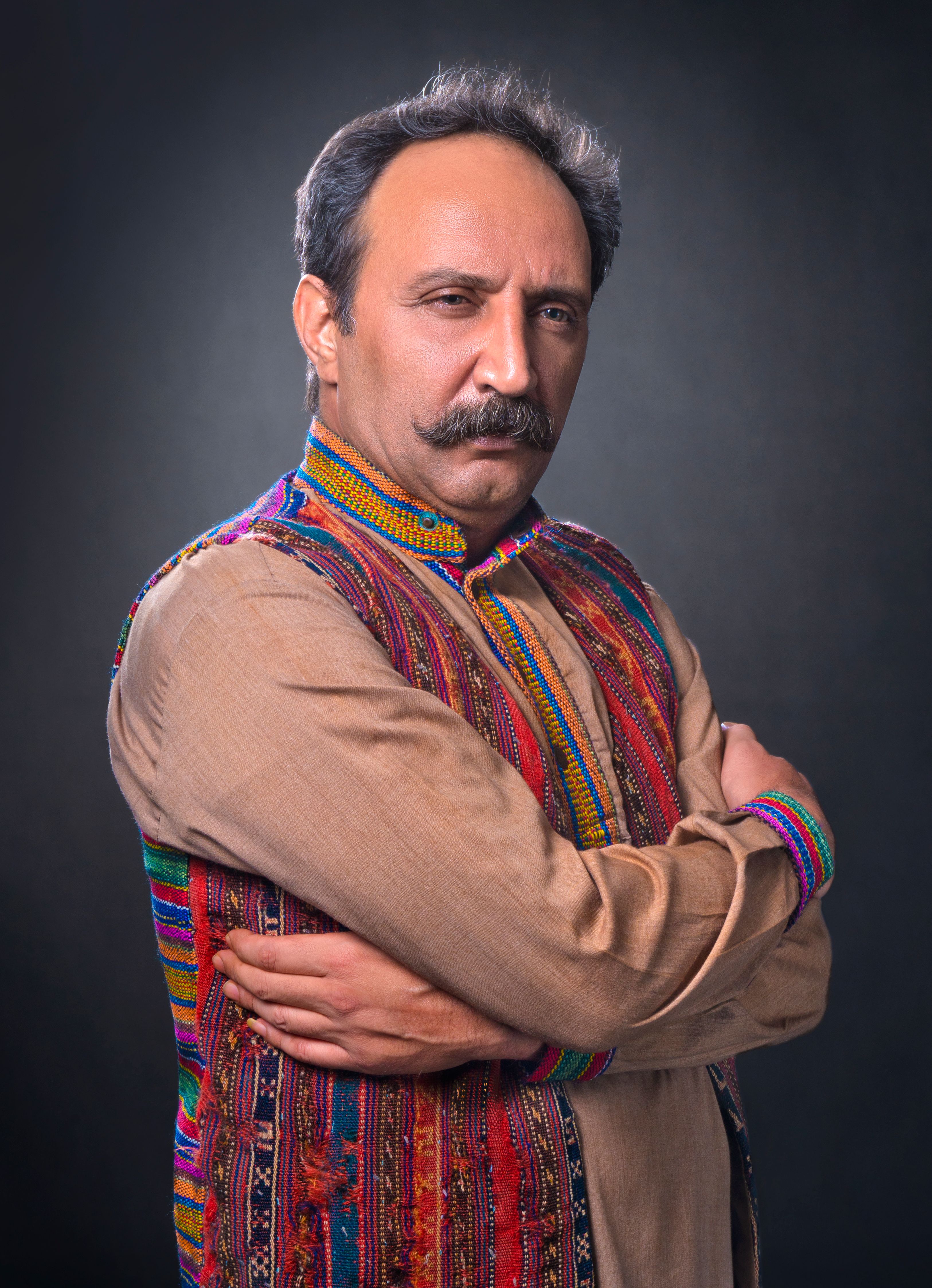 A middle-aged Middle Eastern man with a serious expression poses with crossed arms in traditional attire.