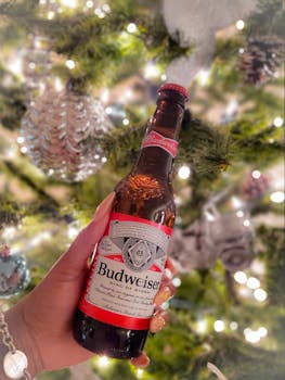 Close-up of a hand holding a Budweiser bottle with a decorated Christmas tree backdrop.