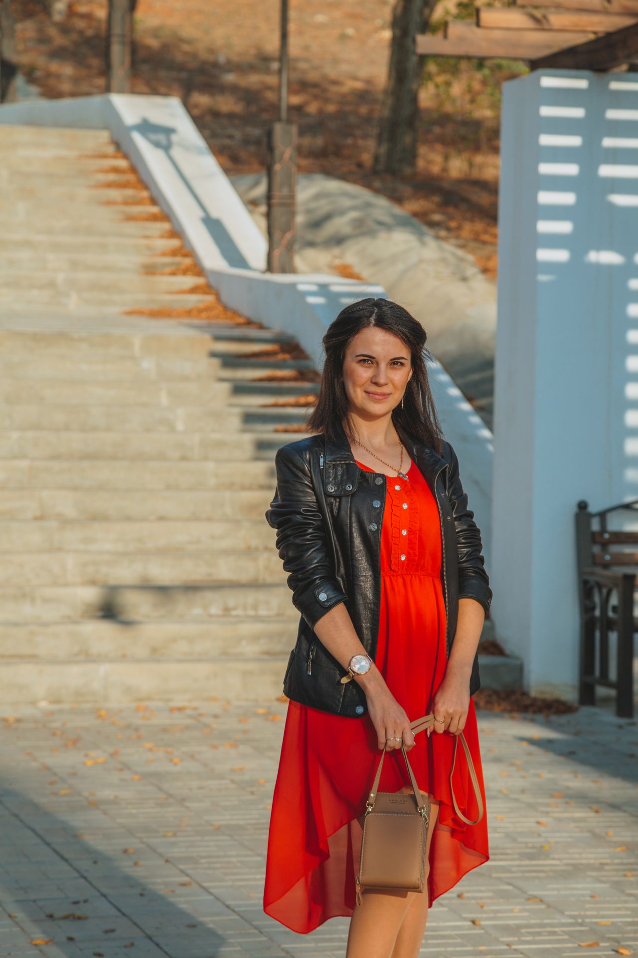 Woman wearing a red dress with a black leather jacket and holding a tan handbag on city steps