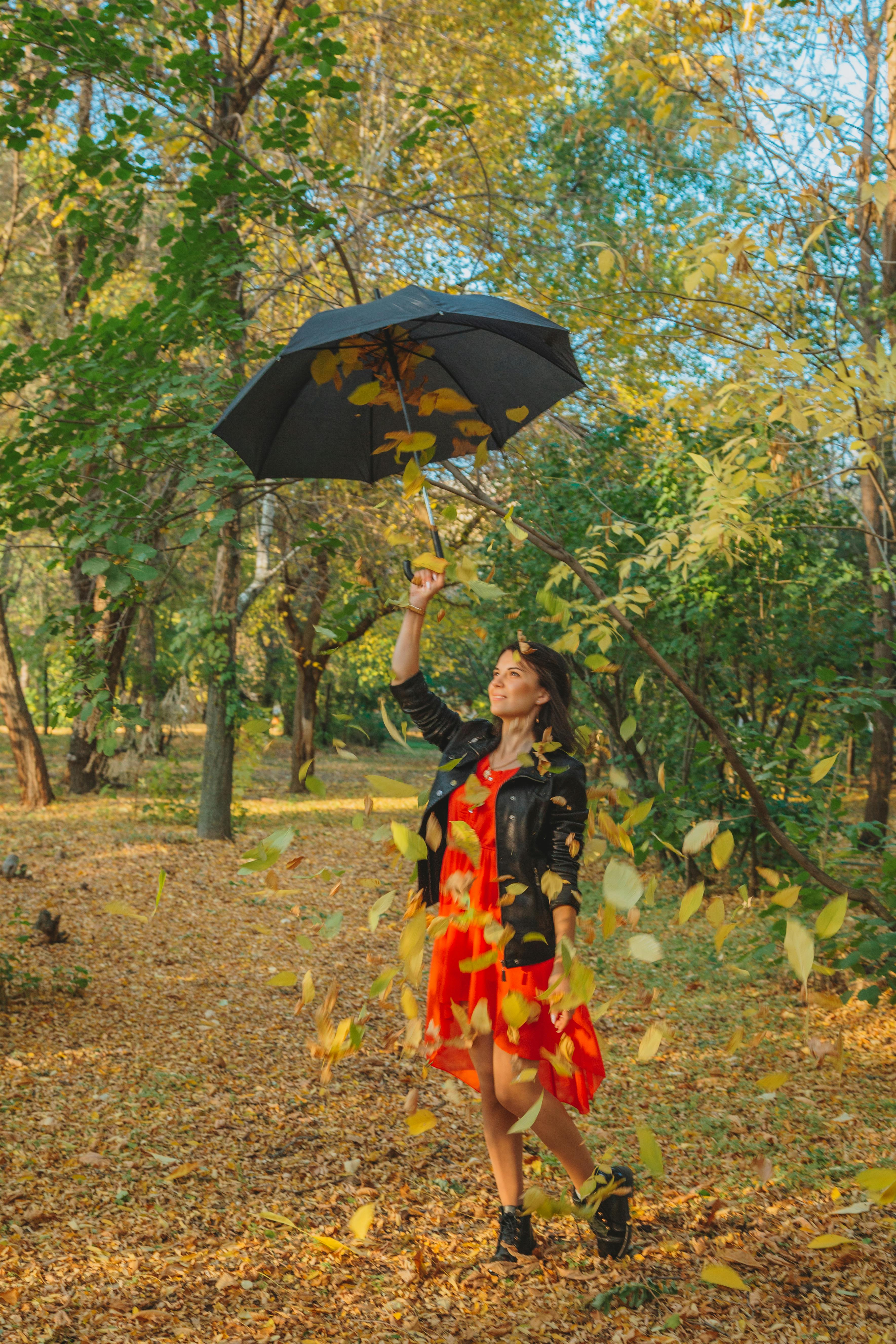Full length of cheerful female in stylish outfit raising umbrella with golden foliage in autumn countryside