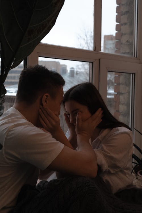 Side view romantic young couple wearing casual clothes sitting together on windowsill and touching each others face gently