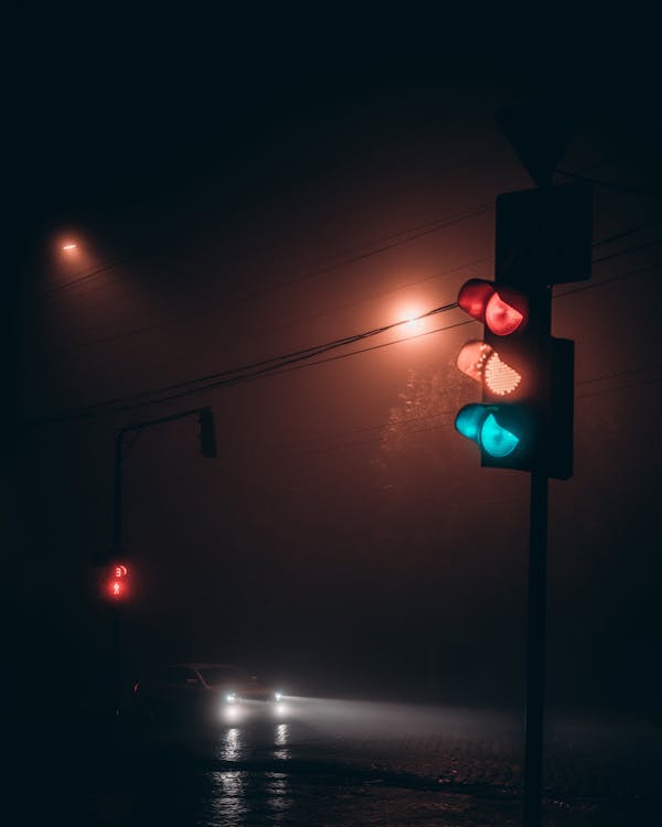 Automobile with glowing headlights on road near illuminated traffic light at foggy night