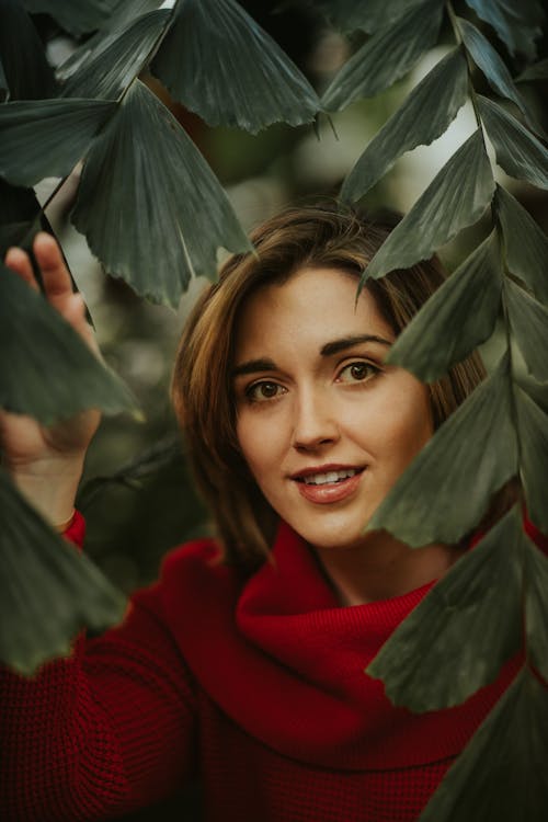 Content female looking at camera through branches with green leaves of exotic plant while standing in garden on blurred background