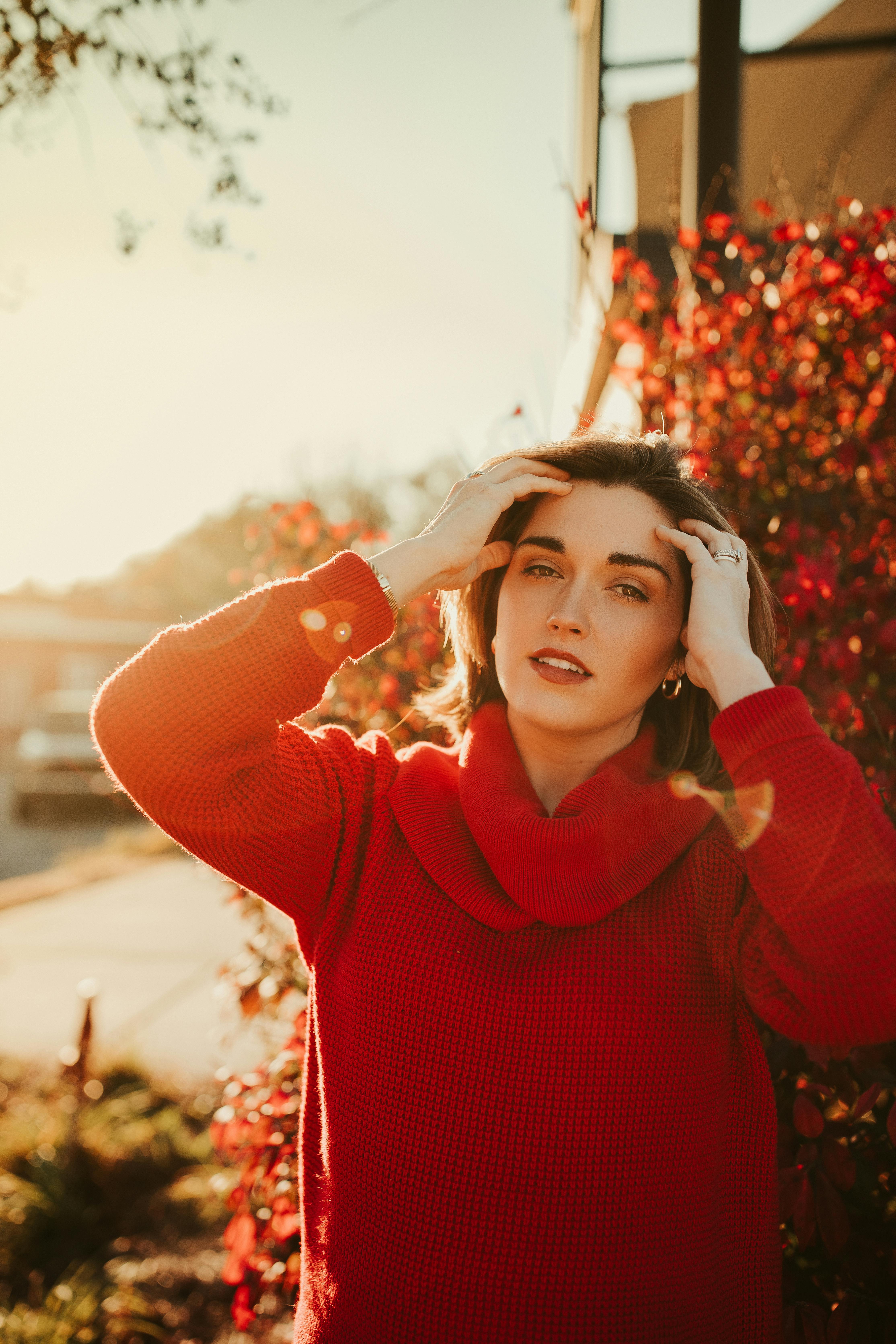 Content female wearing red sweater with hands on head looking at camera while standing on sunny street with plants and buildings on blurred background
