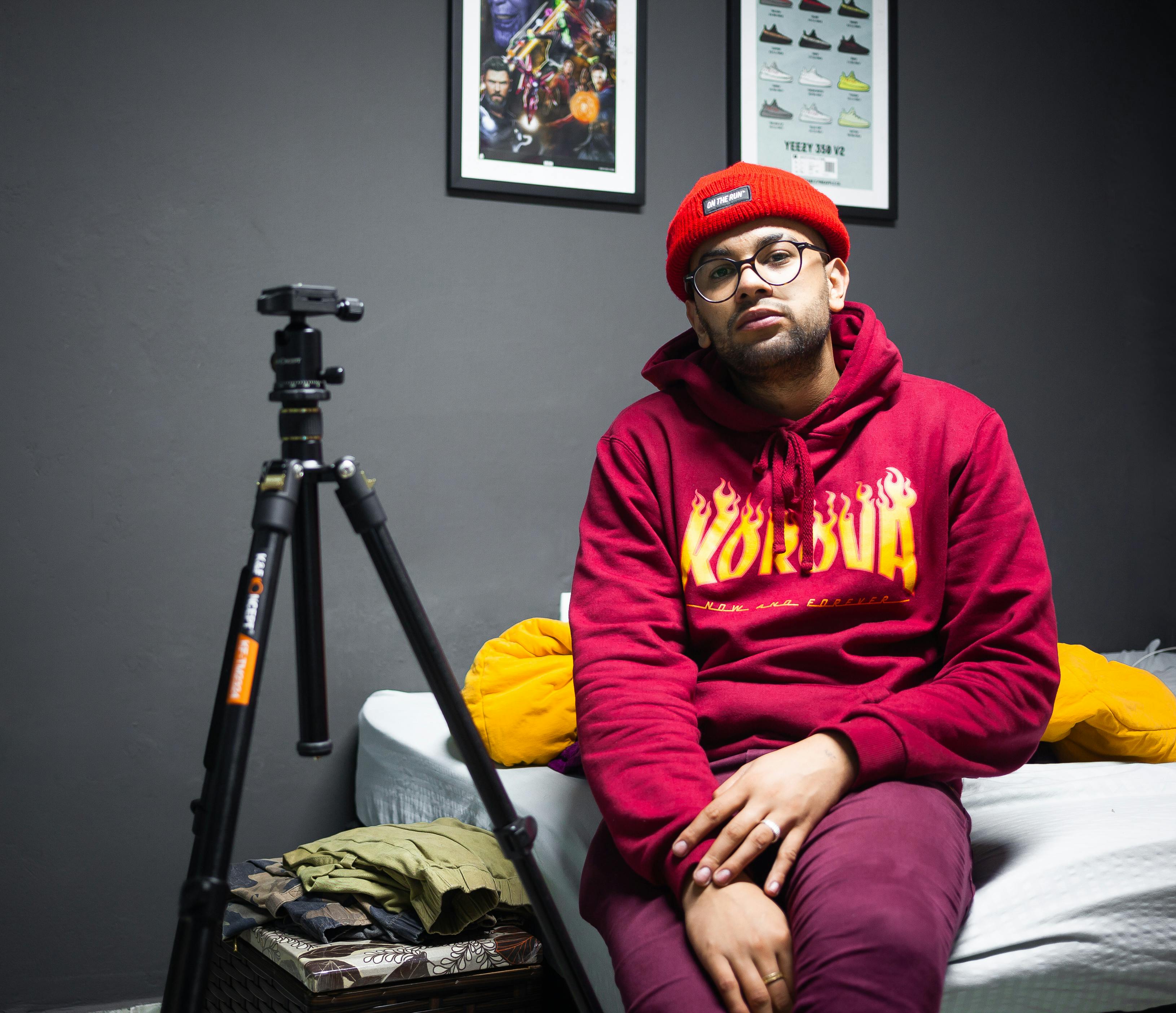 Man in a red hoodie and beanie sitting indoors with a tripod, looking serious.