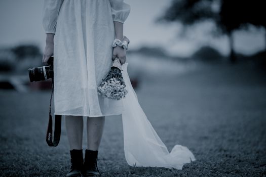 A woman in a white dress stands outdoors holding a camera and bouquet, creating an eerie and serene mood.