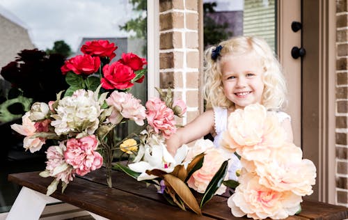 Smiling child in white clothes looking at camera while spending time on terrace at table near house with bouquet of flowers in daytime