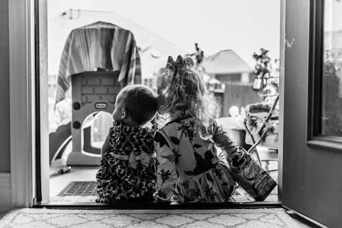 Black and white back view of anonymous kids in dress sitting together in doorway near house with buildings on background in daytime