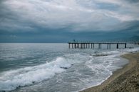 Scenery of endless blue sea waving on sandy beach with wooden pier beneath dramatic cloudy sky on overcast stormy weather