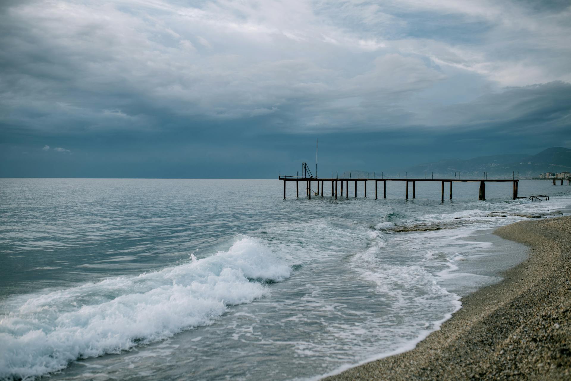 Scenery of endless blue sea waving on sandy beach with wooden pier beneath dramatic cloudy sky on overcast stormy weather