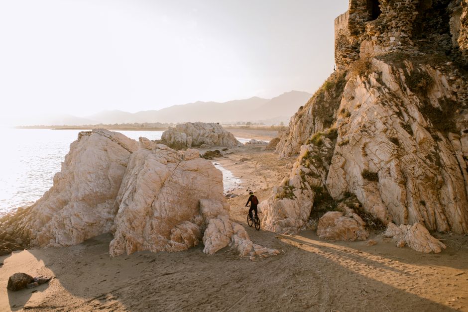 A cyclist navigates rocky coastal terrain by the beach during a picturesque sunset.