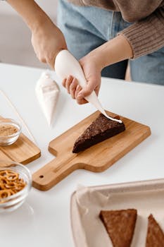 A person decorates a brownie slice with whipped cream on a wooden board.