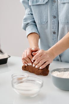 Person kneading chocolate dough with flour and sugar bowls nearby in a kitchen setting.